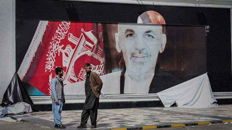 Những người đàn ông Afghanistan đứng cạnh một tấm áp phích rách nát có hình ảnh cựu Tổng thống Afghanistan Ashraf Ghani tại sân bay Kabul, ngày 16/8/2021. Ảnh: AFP